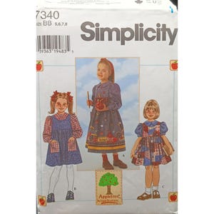 Op de afbeelding: Simplicity naaipatroon voor meisjesjurken. Het patroon toont een tuinbroek, een jurk met lange mouwen met schoolbus- en appelprints en een patchworkjurk. De patroongrootte is BB 5, 6, 7 en 8.