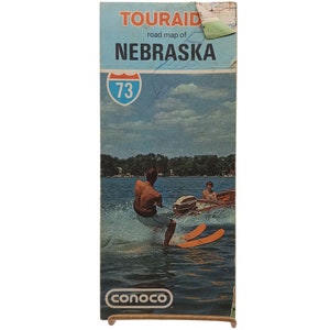 May include: Vintage "Touraid" road map of Nebraska, featuring a waterskiing scene. The map cover is blue with the text "Nebraska" and "73" in a white and red shield. The Conoco logo is at the bottom.