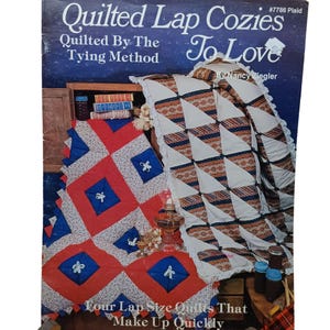 May include: A book cover for a quilting pattern titled "Quilted Lap Cozies To Love" by Nancy Ziegler. The cover features two quilted lap cozies, one with a red and blue checkered pattern and the other with a white, brown, and blue geometric pattern. The book is titled "Quilted Lap Cozies To Love" and is part of the "Quilted By The Tying Method" series.