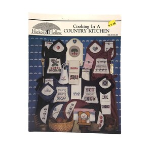 May include: A selection of cross-stitch patterns with a country kitchen theme. The patterns feature aprons, pot holders, towels, and other kitchen accessories with apple, tree, and duck designs. The text "Cooking In A Country Kitchen" is visible.
