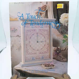 May include: A book titled "A Touch of Tenderness" with a painted clock on the cover. The clock has a light blue face with a white and light blue checkered pattern. The clock is surrounded by a light pink floral design. The book is a Folk Art and Country Collection Presentation by Sandy James & Kay Qnist.