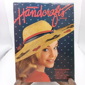 Puede incluir: Una portada de revista con una mujer que lleva un gran sombrero de paja con detalles rojos y negros. La revista se titula "Country Handcrafts Bazaar" y presenta el texto "Patrones de tamaño completo para más de 60 ideas de Bazaar. PLUS sombreros con plantillas, mantas, una muñeca del 4 de julio, modas de verano en punto y ganchillo... y mucho, mucho más."