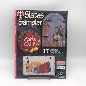 Könnte beinhalten: Ein Buchcover für ein Bastelbuch mit dem Titel "Slates Sampler" mit 17 farbenfrohen bemalten Projekten. Das Cover zeigt ein bemaltes Schieferschild mit der Aufschrift "Mom's Cafe" und einen bemalten Schieferengel mit einem roten Kleid und gelben Blumen.