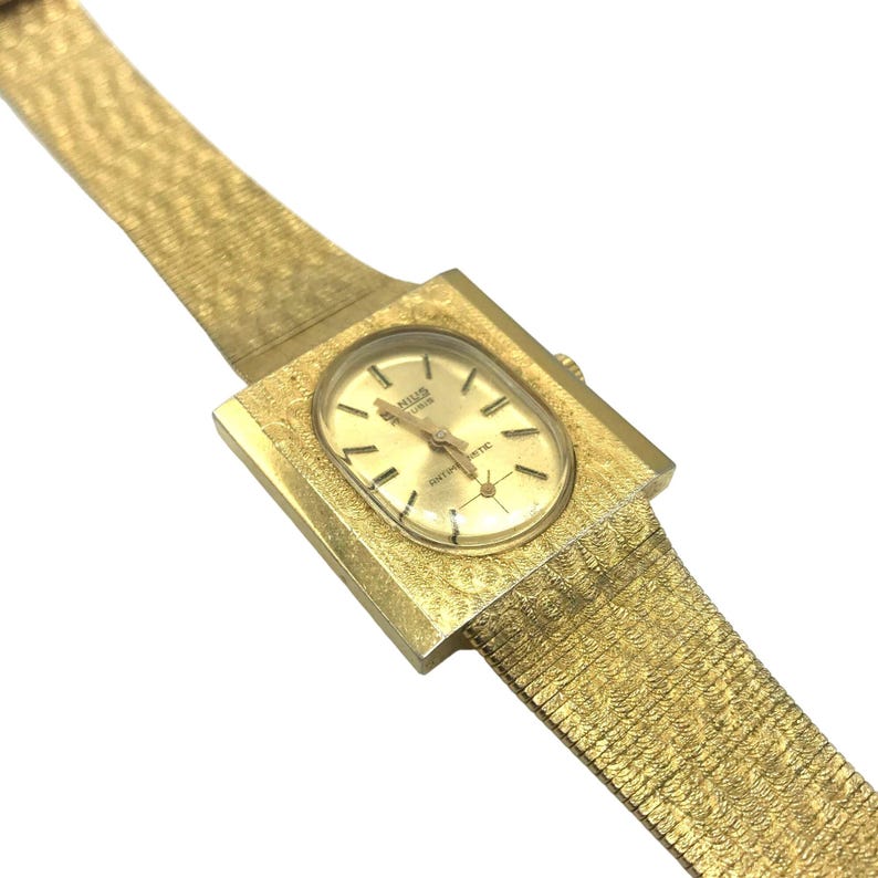 Puede incluir: Un reloj de pulsera dorado con una caja cuadrada y una banda dorada texturizada. La esfera del reloj es de forma ovalada y presenta el texto "KIESLER GENEVA AUTOMATIC".