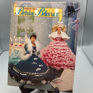 May include: A crochet pattern book for making birthday doll dresses. The cover features two dolls dressed in pink and blue crochet dresses with crowns. The book is titled "Birthday Belles Vol. II" and is published by Annie's Attic.