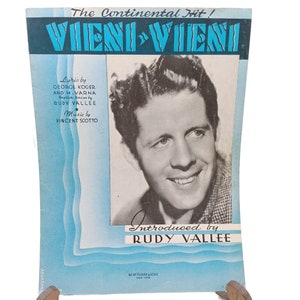 Puede incluir: Una portada de partitura musical vintage con una foto en blanco y negro de Rudy Vallee, un cantante popular de la década de 1930. La portada es azul con olas blancas y el título "VIENI VIENI" en letras grandes y negritas. El texto "The Continental Hit!" está impreso sobre el título.
