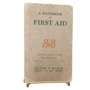 May include: A vintage first aid handbook with the title "A Handbook of First Aid" in green lettering. The cover is tan with the logo "BAB" in orange. The book is propped up on a gold-coloured stand.