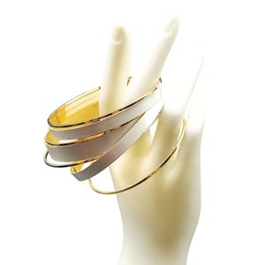 Könnte beinhalten: Gold- und weiß emaillierte Armreifen. Das Bild zeigt goldfarbene Metallarmreifen, einige mit weißer Emaille-Beschichtung. Die Armreifen werden auf einem Mannequin-Hand präsentiert, um ihr Design zu zeigen. Ein klassisches Schmuckstück.