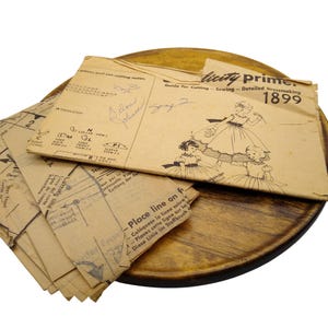 May include: Vintage dressmaking pattern from 1899, featuring illustrations of women's dresses. The pattern is printed on aged, tan paper and includes handwritten notes. The pattern is placed on a round, wooden tray.