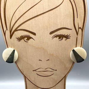 May include: A pair of round earrings with a cream-colored background and a dark gray triangle design. The earrings are displayed on a wooden face-shaped stand.