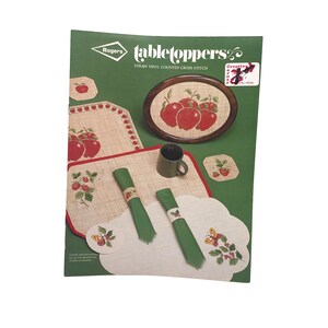 May include: A vintage craft pattern booklet for "Tabletoppers" straw vinyl counted cross stitch featuring a red apple design. The cover shows a variety of finished projects including a tray, placemats, coasters, and napkin rings.