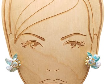 May include: A pair of clip-on earrings with a floral design. Each earring features light blue and clear crystals, with a gold-colored metal base. The earrings are displayed on a wooden face-shaped jewelry holder.