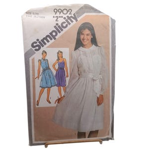 Può includere: Un pacchetto di modelli di cucito Simplicity vintage. Il pacchetto presenta un disegno di due abiti, uno blu e uno viola, e una foto di una donna che indossa un abito bianco. Il pacchetto è etichettato "Simplicity" e include la taglia 9/10.