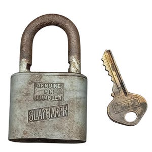Puede incluir: Un candado Slaymaker plateado vintage con una llave a juego. El candado tiene un asa curva y las palabras "Genuine Pin Tumbler" y "Slaymaker" están impresas en la parte delantera. La llave es de color dorado claro.