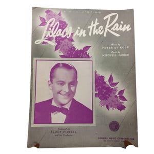 以下が含まれることがあります： 「Lilacs in the Rain」の楽譜の表紙。テディ・パウエルの肖像画が特徴です。表紙は、紫色の花のアクセントとテキストが施された灰色の背景です。テキストには、曲名、作曲者、作詞家、出版社が含まれています。