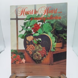 Pode incluir: Um livro intitulado "Heart to Heart... a country collection" de Gretchen Cagle. A capa apresenta um vaso de madeira pintado em forma de coração com uma planta de gerânio dentro. O vaso está sentado em uma bandeja de madeira.