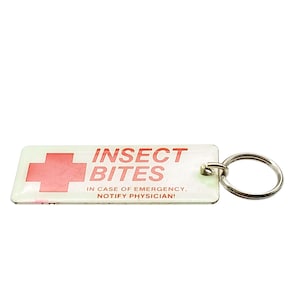 May include: A rectangular white keychain featuring a red cross symbol and the red text INSECT BITES. Below this, smaller red text reads IN CASE OF EMERGENCY, NOTIFY PHYSICIAN! The keychain includes a silver metal ring for attachment.