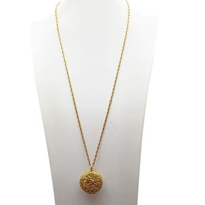 May include: A gold-colored necklace with a long chain and a round pendant. The pendant features an intricate, detailed design. The necklace is displayed on a white mannequin, showcasing the jewelry's design and color.