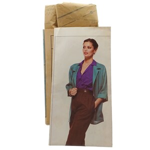 May include: A teal blazer with a purple blouse and brown pants. The model is wearing a gold bracelet and a silver necklace.