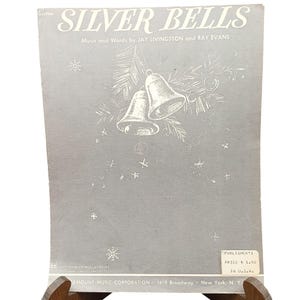 Op de afbeelding: Een vintage bladmuziekomslag voor Silver Bells door Jay Livingston en Ray Evans. De omslag is lichtgrijs met witte lijntekeningen van twee klokken, dennentakken en sterren. De tekst bevat de titel, de auteurs en uitgeversinformatie.