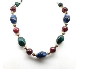 Puede incluir: Un collar colorido con grandes cuentas pulidas en tonos azul, verde y burdeos. Los espaciadores dorados más pequeños están intercalados. El collar se exhibe en un busto de maniquí blanco, mostrando su diseño y paleta de colores.