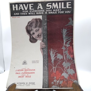 Puede incluir: Una portada de partitura musical vintage con una mujer asomándose desde detrás de un diseño floral rojo y blanco. El título de la canción es "Have a Smile" con la letra "For everyone you meet and they will have a smile for you".