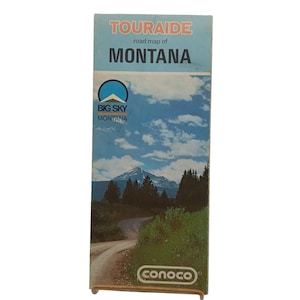 May include: A vintage "TOURAIDE" road map of Montana. The cover features a scenic landscape with a winding road, mountains, and a blue sky with clouds. The "BIG SKY MONTANA" logo is visible, along with the "conoco" logo.