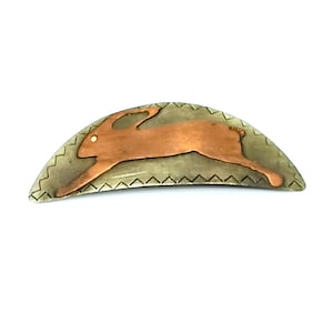 May include: A silver and copper hair clip shaped like a crescent moon with a stylized copper rabbit design.