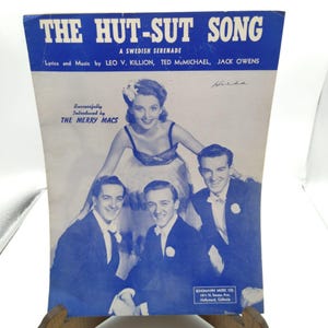Puede incluir: Partitura vintage titulada "THE HUT-SUT SONG", una serenata sueca. La portada azul y blanca presenta una fotografía de una mujer y tres hombres. El texto incluye el título de la canción, el género y los nombres de los letristas y músicos.