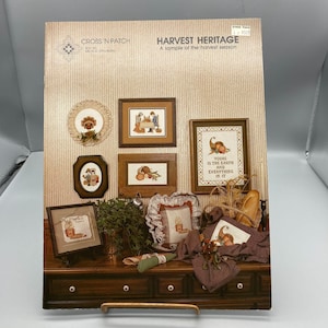 May include: A cross-stitch pattern book titled "Harvest Heritage" with various framed cross-stitch samplers featuring fall harvest themes. The book cover displays the title and the Cross 'N Patch logo. The samplers include pumpkins, cornucopias, and text.