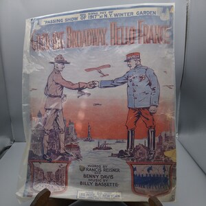May include: A vintage sheet music cover featuring a soldier from the US Army shaking hands with a soldier from the French Army. The cover is titled "Goodbye Broadway, Hello France" and features a cityscape of New York City in the background. The text on the cover reads "Big Song Hit of "Passing Show of 1917" at N.Y. Winter Garden."