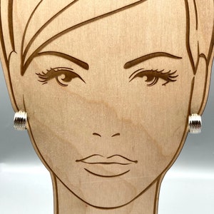 May include: A pair of silver hoop earrings with a textured, ridged design. The earrings are shown on a wooden mannequin head.