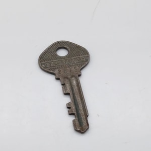 Puede incluir: Una llave de metal vintage con la inscripción Slaymaker 215. La llave tiene una cabeza redondeada con un ojo de cerradura y un borde dentado.