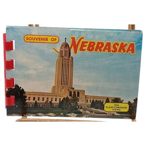 Può includere: Un libretto souvenir vintage intitolato "Souvenir of Nebraska" con una fotografia di un edificio alto sotto un cielo blu. Il libretto ha una rilegatura a spirale rossa e supporti in legno. È visibile anche il testo "TEN PLASTICHROME VIEWS".