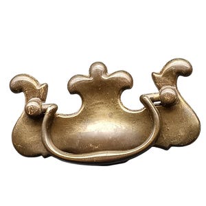 Op de afbeelding: Antieke messing ladeknop met een decoratief ontwerp. De knop heeft een centrale handgreep met een gebogen vorm en twee zij-elementen. Het messing heeft een warme, verouderde patina, wat een vintage stijl suggereert. Ideaal voor meubelrestauratie.