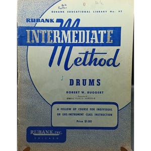 May include: A vintage music instruction book titled Rubank Intermediate Method for Drums by Robert W. Buggert. The cover is off-white with blue text and graphics, featuring the Rubank Educational Library No. 97 branding and O'Neill Public Schools.