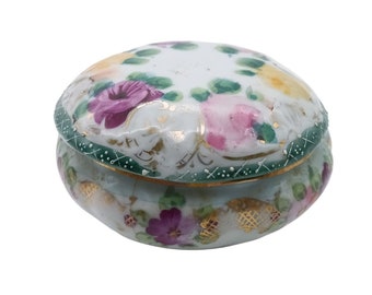 May include: A small, round, white porcelain trinket box with a lid. The box is decorated with hand-painted floral designs in shades of pink, purple, and green. The lid has a gold trim and the box has a green trim.