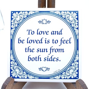 Könnte beinhalten: Eine blaue und weiße Keramikfliese mit einem dekorativen Rand und einem Zitat, das lautet: "Liebe zu geben und geliebt zu werden, bedeutet, die Sonne von beiden Seiten zu spüren." Die Fliese ist mit 1997 datiert.