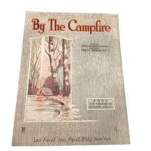 Pode incluir: Capa de partitura musical vintage intitulada "By The Campfire". A capa apresenta uma ilustração de uma cena de acampamento com árvores, um rio e uma tenda. O texto inclui informações sobre o letrista, compositor e editor.