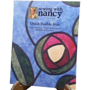 May include: A sewing book titled "sewing with nancy" by Nancy Zieman. The cover features a floral design in shades of red, green, and yellow on a blue background. The book is propped up on a wooden stand.