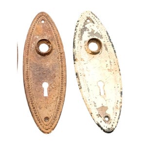 May include: Two antique oval-shaped door plates. One is a tarnished bronze color with a keyhole and decorative border. The other is painted white with chipped paint, also with a keyhole and decorative border. Both have a circular hole.