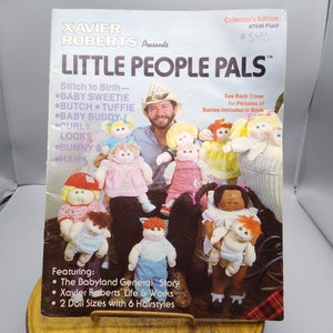 May include: A book cover featuring a photo of a man with several dolls. The book title is "Little People Pals" and the subtitle is "Stitch to Birth". The book cover also includes a list of doll names, such as "Baby Sweetie", "Butch", "Tuffie", "Baby Buddy", "Curly Locks", "Bunny & Hank". The book cover also includes the text "The Babyland General Story", "Xavier Roberts' Life & Works", and "2 Doll Sizes with 6 Hairstyles".