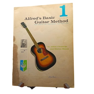 May include: A vintage book cover for "Alfred's Basic Guitar Method." The cover features a drawing of an acoustic guitar in shades of brown, orange, and black. The number "1" is in a bright blue box. The book is for group or individual instruction.