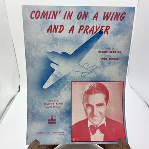 May include: A vintage sheet music cover for the song "Comin' In On A Wing And A Prayer" featuring a blue and white aeroplane illustration and a photo of Henry King, the song's performer. The text "Lyric by Harold Adamson, Music by Jimmy McHugh" is printed on the cover.