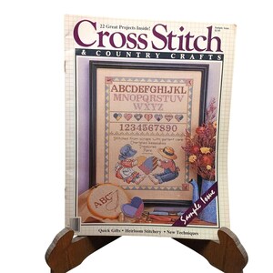 Könnte beinhalten: Eine Musterausgabe des Magazins Cross Stitch & Country Crafts mit einem gerahmten Sampler, der ein Alphabet, Zahlen und zwei Kinder mit einem Herz zeigt. Das Cover enthält einen kleinen Stickrahmen mit den Buchstaben ABC und ein Trockenblumenarrangement.