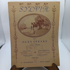 May include: A vintage sheet music cover for the song "Oley Speaks" by Sylvia. The cover features a romantic illustration of a couple standing under a tree in a landscape setting. The text "Oley Speaks" is prominently displayed in a decorative font.