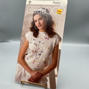 May include: A white knitted top with a cable knit design and colorful pom-poms. The top has short sleeves and a round neckline. The pattern is called "Poppy" and the style number is 739. The price is $1.50.