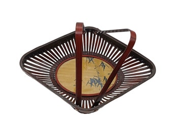 Può includere: Un cesto quadrato in bambù marrone con un design intrecciato e una base rotonda decorativa gialla e rossa con illustrazioni di bambù nero. Il cesto ha due manici marroni.