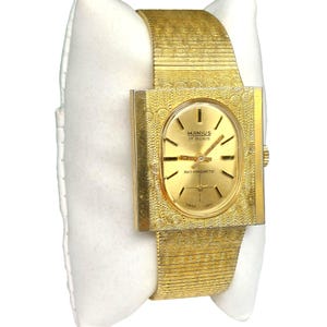 Puede incluir: Un reloj de pulsera dorado con una esfera cuadrada y una correa de metal dorado. La esfera del reloj es de forma ovalada y tiene un fondo blanco con marcas negras. La esfera del reloj tambi&eacute;n tiene el texto "MANIUS 17 RUBIS" y "ANTIMAGNETIC".