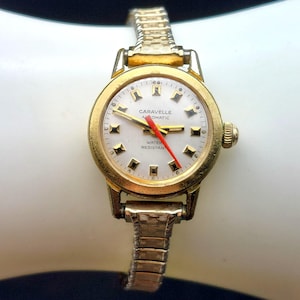 May include: A gold-toned Caravelle automatic wristwatch with a white face and red second hand. The watch features a gold bezel and a flexible gold band. The watch face displays the words "Caravelle Automatic" and "Water Resistant".